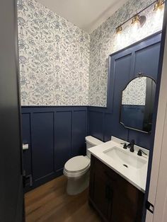 a white toilet sitting under a bathroom mirror next to a wooden sink vanity and blue wallpaper