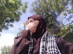 a woman wearing a brown hijab covering her face with her hands while standing in front of some trees