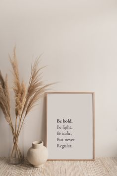 a white vase sitting next to a plant on top of a wooden table in front of a framed poster