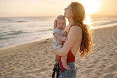 a woman holding a baby on the beach at sunset