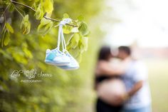 a couple cuddles under a tree with their baby shoes hanging from the branch