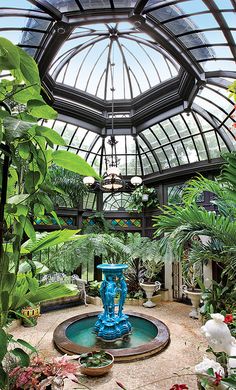 the inside of a glass house with plants and water fountain in the center, surrounded by greenery