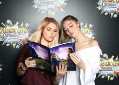 two beautiful women standing next to each other holding an open book