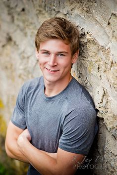 a young man leaning against a stone wall with his arms crossed and looking at the camera