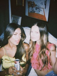 two young women sitting on a bed looking at their cell phones and drinking sodas