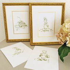 two framed pictures sitting on top of a table next to flowers and watercolor pencils