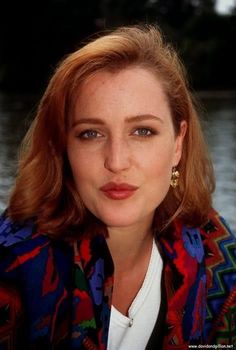 a woman with red hair and blue eyes wearing a colorful jacket in front of water