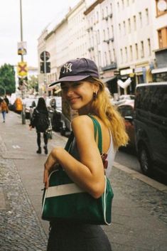 a woman is walking down the street carrying a purse and smiling at the camera while wearing a baseball cap