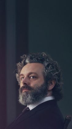 a man with curly hair and beard wearing a suit looking off into the distance while sitting in a chair