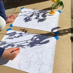 two people are drawing on paper with blue tape and some flowers in the back ground