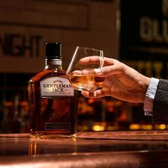 a person holding a glass next to a bottle of whiskey on a bar counter top