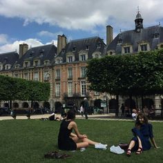 two women are sitting on the grass in front of a large building with many windows