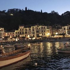 several boats are docked in the water at night near some buildings and lights on top of a hill