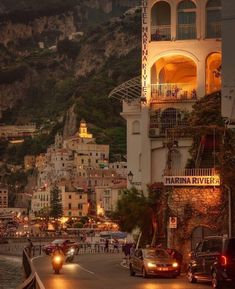 cars are driving down the road in front of some buildings and cliffs at night time