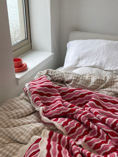 an unmade bed with a red and white blanket on top of it next to a window