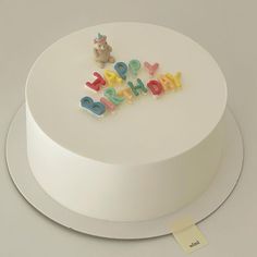 a white cake with the word happy birthday spelled out in colorful letters on top and a teddy bear figurine next to it