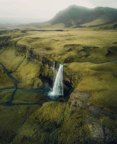 an aerial view of a waterfall in the middle of a green field with mountains behind it