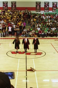 two girls in graduation gowns stand on the basketball court as people watch from the stands