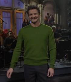 a man in a green sweater standing on a stage with other people behind him and an audience