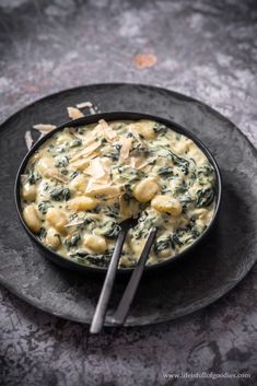 a black plate topped with pasta and spinach covered in sauce next to two silver spoons