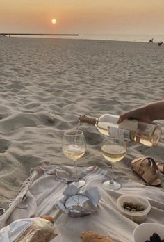 two people are sitting on the beach with wine and bread in front of them, while one person is pouring white wine into a glass