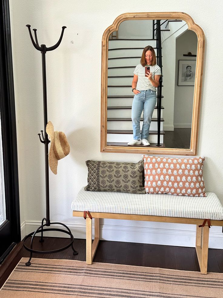 a woman taking a selfie in the mirror while sitting on a bench next to a hat rack