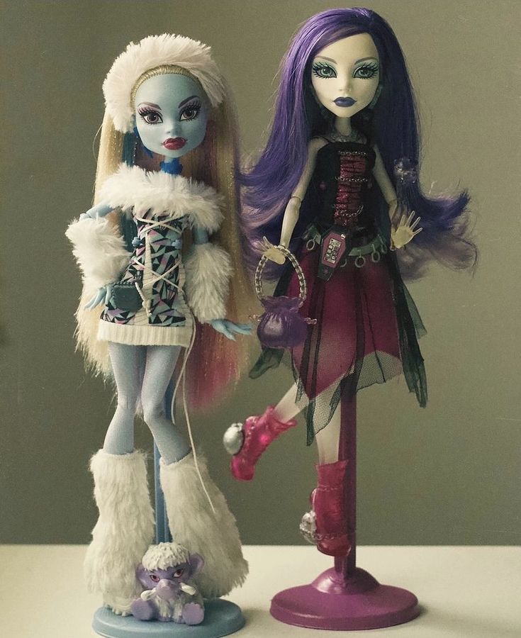 two dolls standing next to each other on top of a white table with purple hair