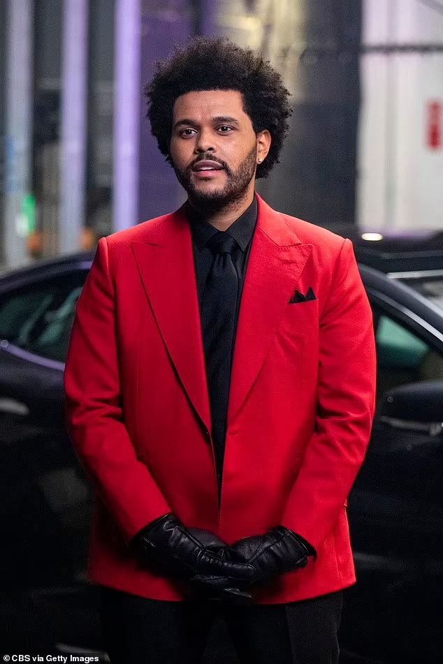 a man in a red suit and black tie standing next to a car with his hands on his hips