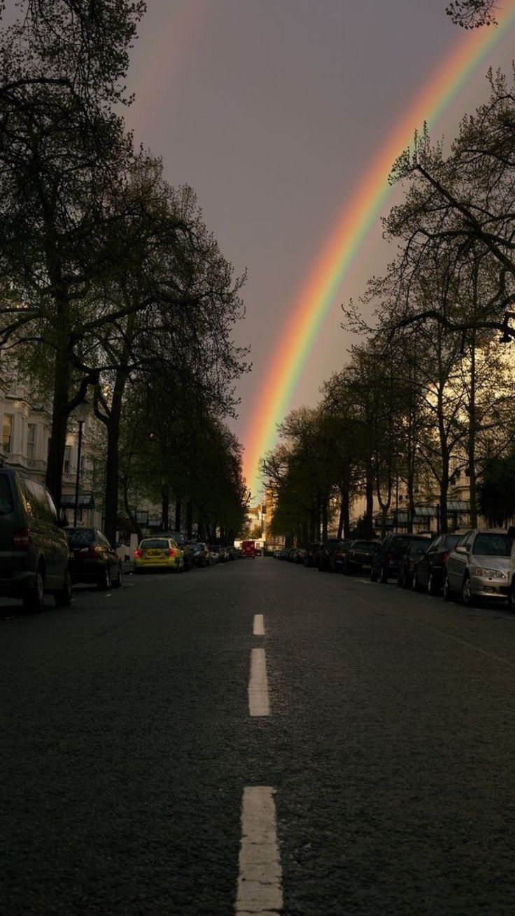 two rainbows are seen in the sky over a street lined with parked cars and trees