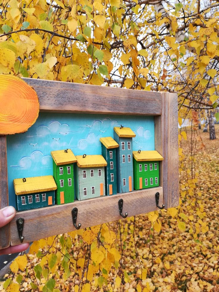 a person holding up a wooden frame with small houses on it in the fall leaves