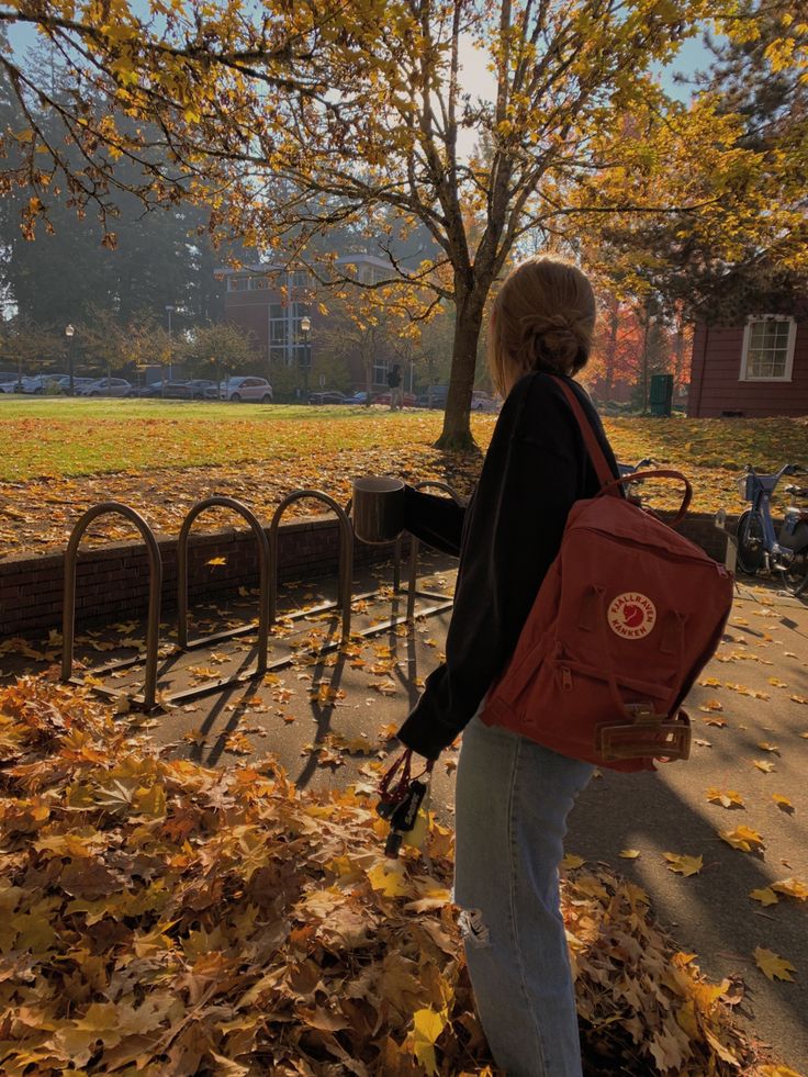 a woman with a red backpack is walking in the leaves near a fence and trees