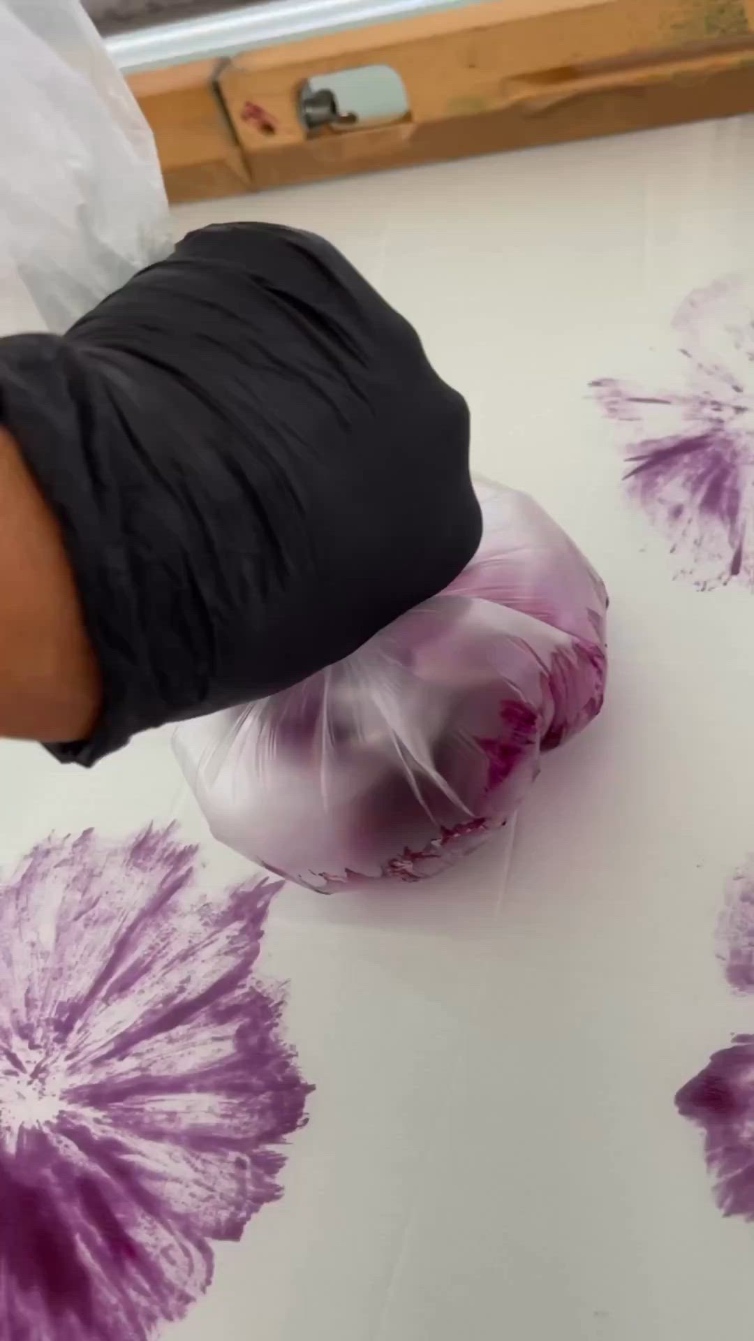 This may contain: a person is painting with purple flowers on a white table cloth and black gloves are holding the fabric