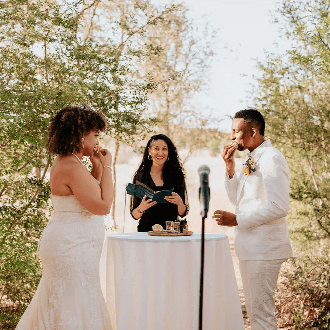 a couple in the foreground wearing all white and their officiant standing behind a table in all black, during the Tasting the Four Elements wedding ceremony ritual