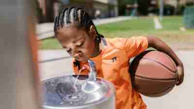 A child drinking water from a fountain while holding a basketball. Summer hydration tips for children with ADHD, autism.