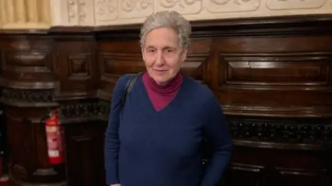 Jenny Jenny, with short grey hair, is looking straight at the camera in a wood-panelled room 