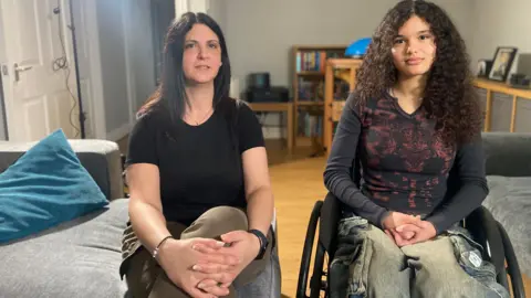 Christina sits in a chair in a living room next to her daughter Mia who is seated in a wheelchair