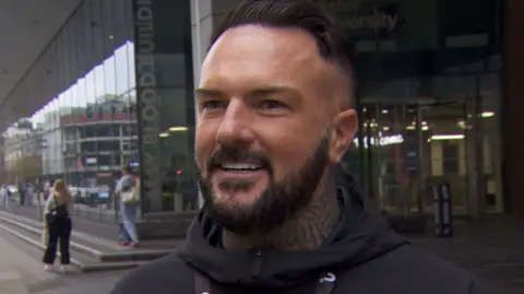 A man with a black beard and black hair stands in front of University of Ulster building wearing a black hoody