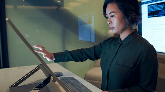 Страничен профил на жена, носеща тъмна риза в тъмен офис, преглеждаща или работеща на Microsoft Surface Studio.