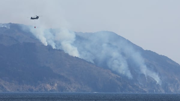 A helicopter is pictured as smoke rises due to a wildfire on a mountainside near the city of Ofunato