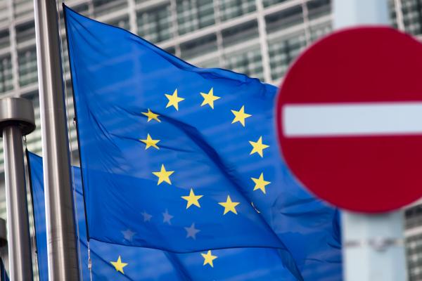 The image features a European Union flag with its circle of twelve golden stars on a blue background, waving in front of a modern glass building. In the foreground is a blurred 'no entry' traffic sign, against the clearly focused EU flag.