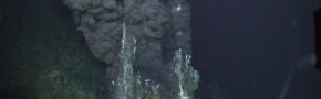 An underwater photo with a blue backdrop and a mineral formation erupting black, mineral-rich fluid.