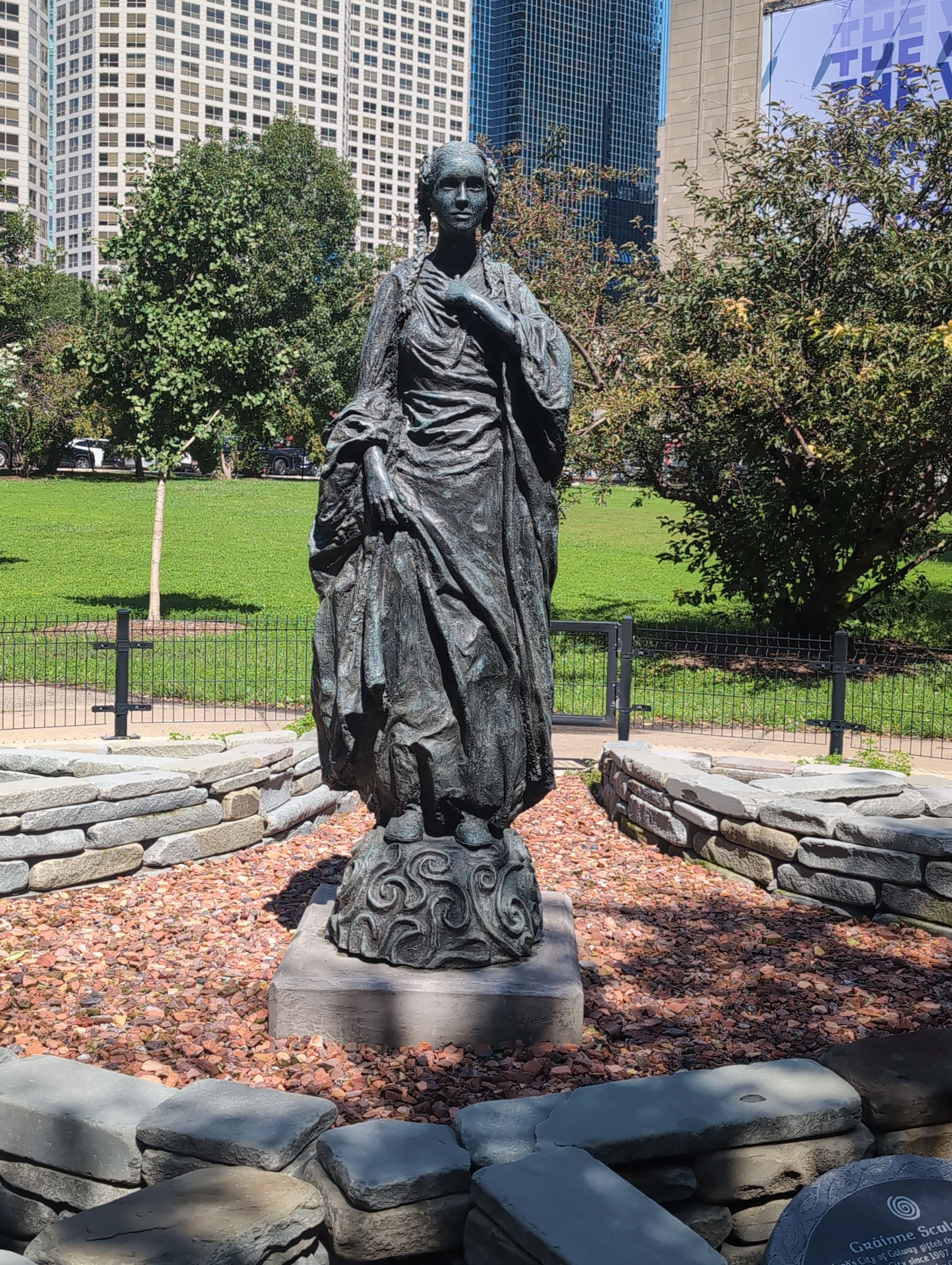 A statue of a woman with braided hair and long flowing robes atop a mound covered in swirl patterns. It sits in a park on red rocks surrounded by bricks. Grass and trees can be seen beyond it, and further back, tall buildings.