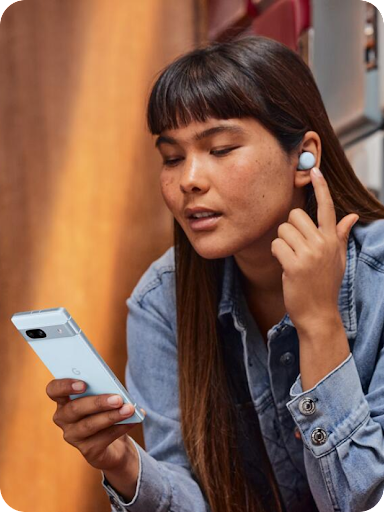 An Android user is holding a conversation, touching their earbud with their finger and looking down at their Android phone.