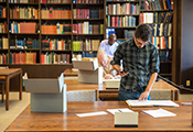 two students standing at desks with floor-to-ceiling bookcase behind them