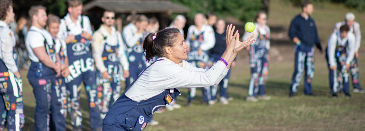 Kvinna i blå hängselbyxor med märken fångar boll utomhus. I bakgrunden fler personer som är likadant klädda.