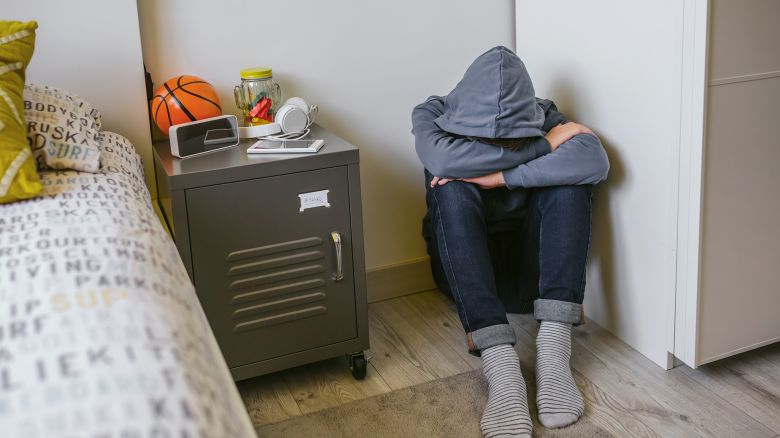 Unrecognizable teenager boy with anxiety sitting on the floor of his bedroom with his head resting on his arms