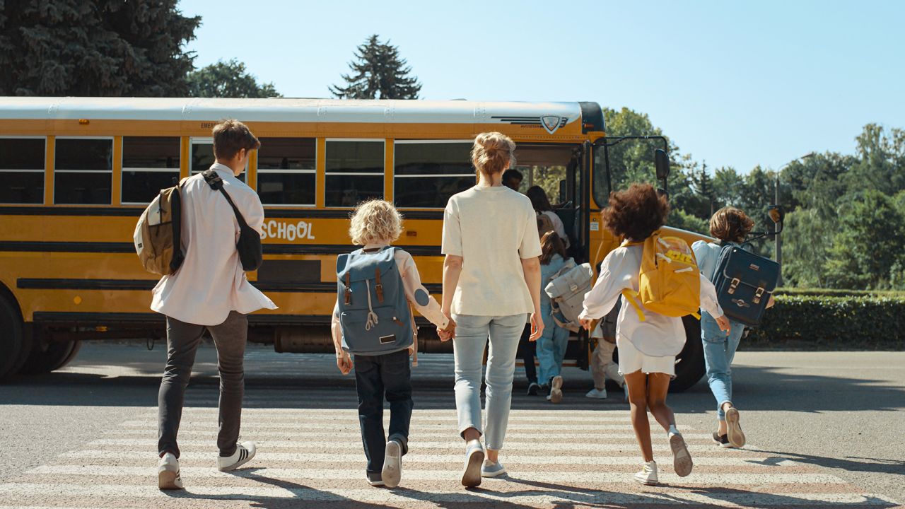 Mother hold hand schoolboy walking to yellow schoolbus. Energetic teenage pupils walking crossway. Young mom accompany curly son to school sunny morning. Teens crowd running boarding academic shuttle.