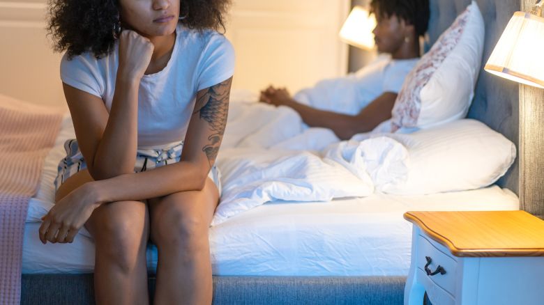 A woman is feeling lonely sitting on the edge of a double bed by herself, leaning her head on her hands and her elbow on her knees, as her husband is sitting up in bed on the other side of the bedroom