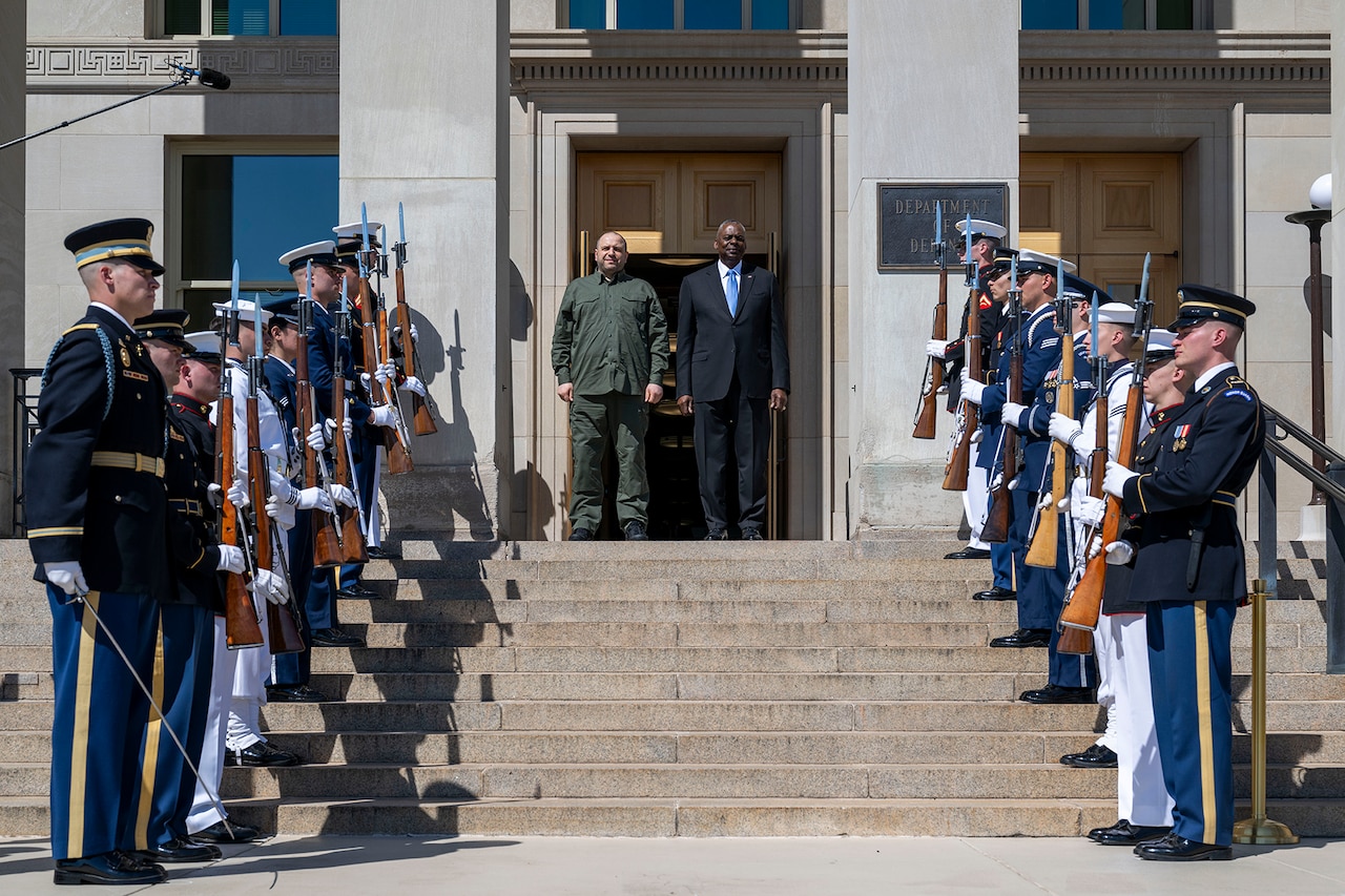Two men stand next to one another at the top of some stairs outside a building. Service members line the stairs from bottom to top.
