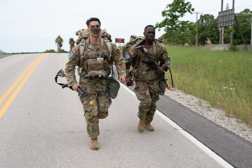 Two soldiers run downhill in gear ahead of a pack on an American road in the countryside.